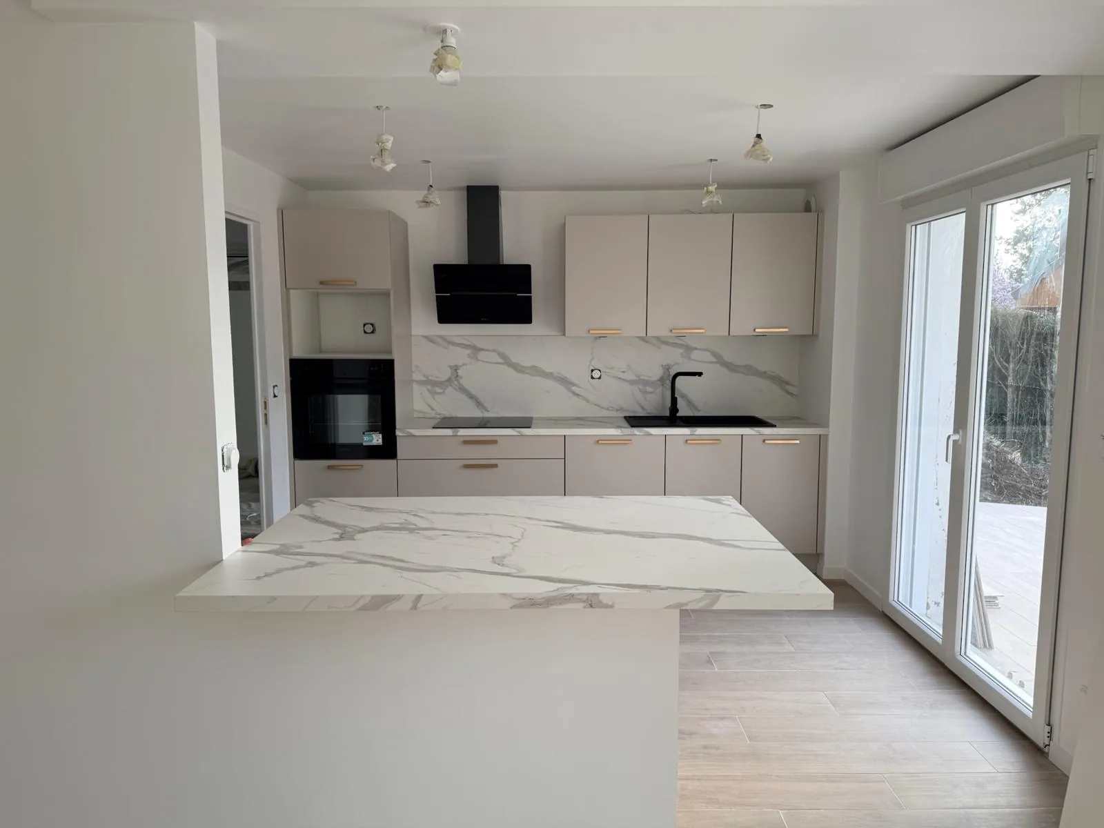 Cuisine ouverte rénovée dans un pavillon : façades beige, poignées dorées, crédence et îlot effet marbre, hotte noire