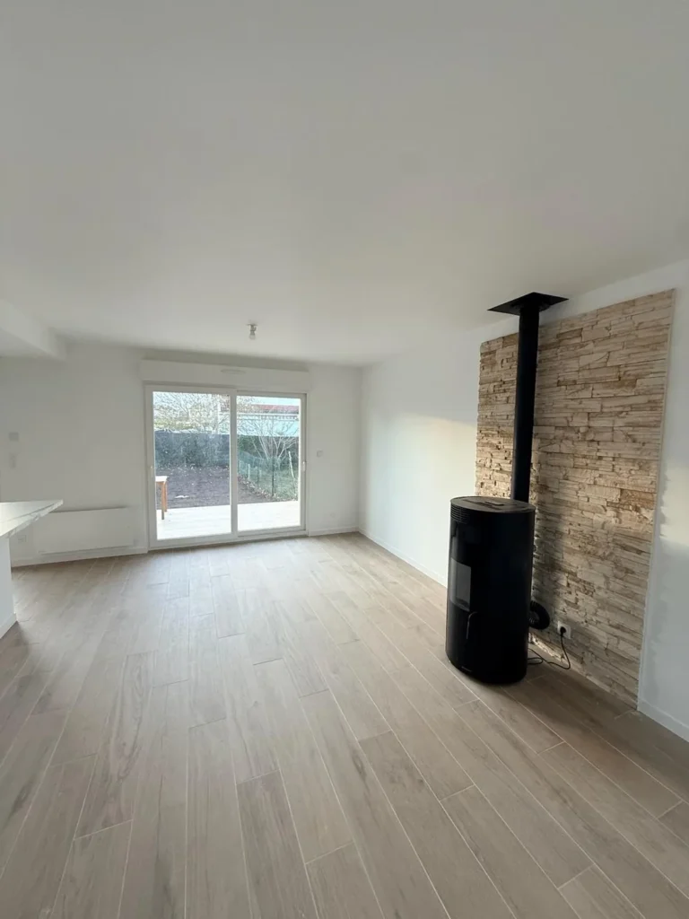 Séjour rénové dans un pavillon : murs blancs, carrelage effet bois, poêle noir et parement pierre, baie vitrée sur le jardin